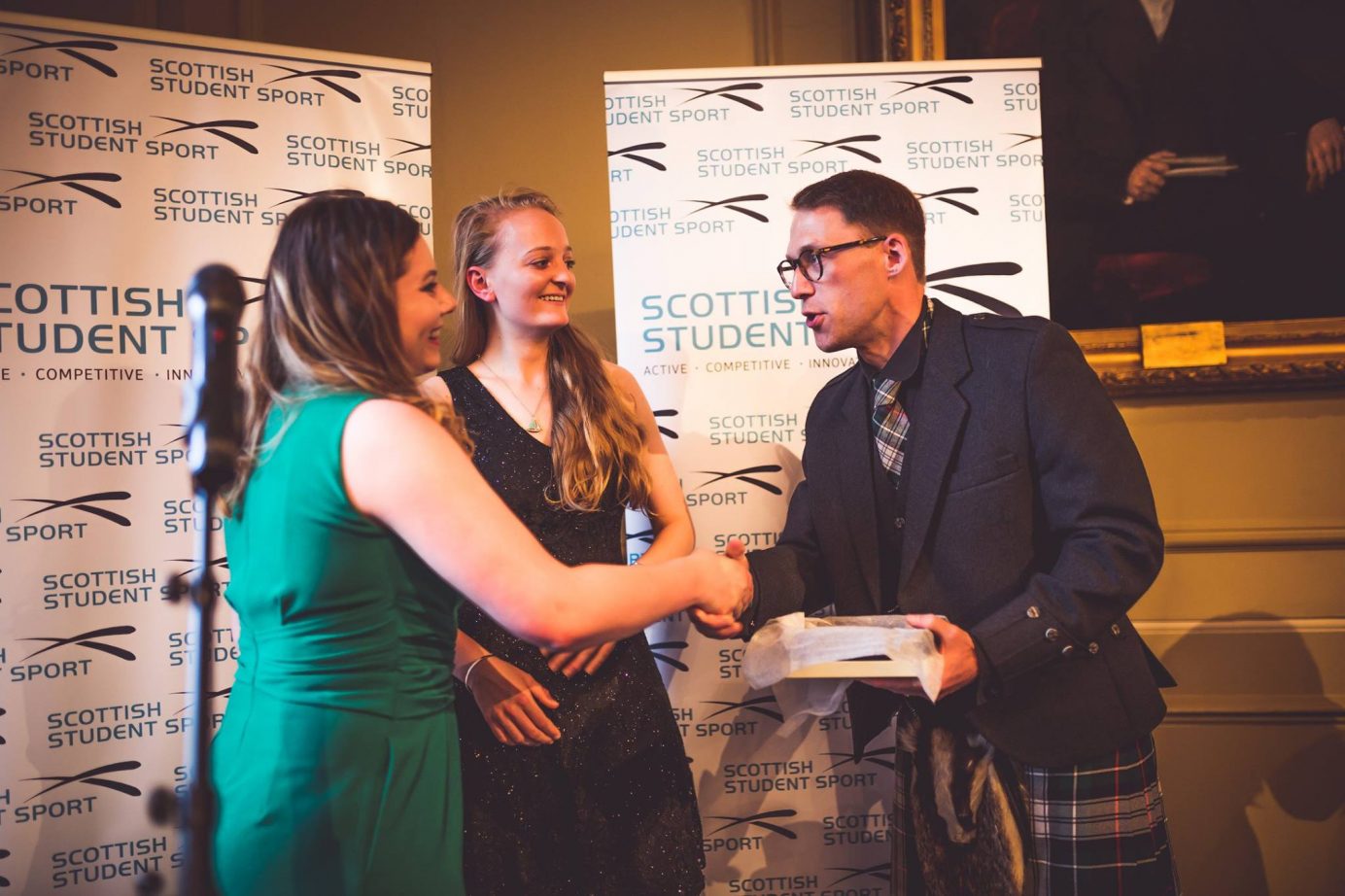 Scottish Student Sport Chair Pete Burgon awarding two female students with a box and shaking their hand. Behind them are two white Scottish Student Sport pull up banners with blue writing